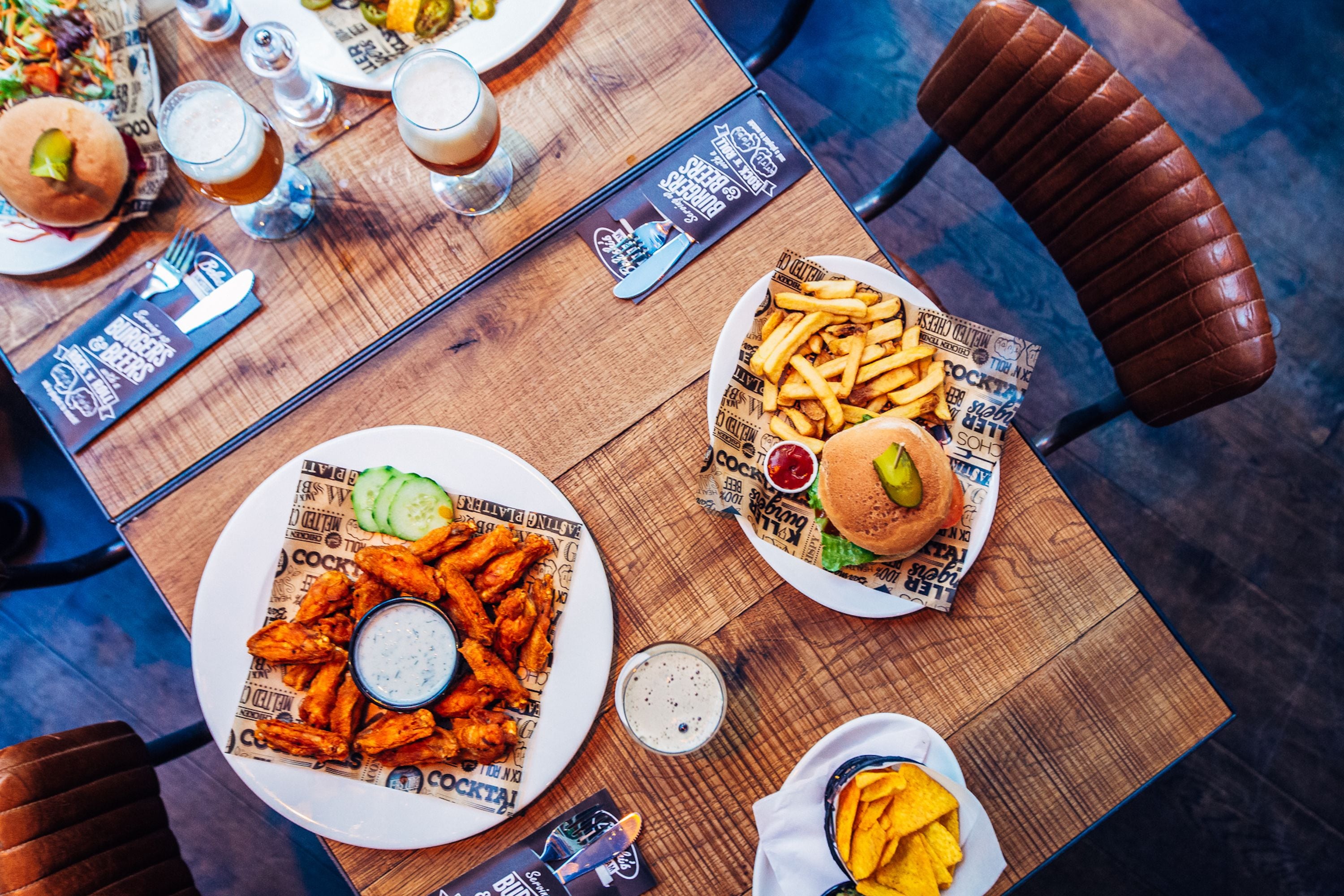 Et spisebord med tallerkener med mad, blandt andet burgere, pommes frites og kyllingevinger, samt drikkevarer og tilbehør.