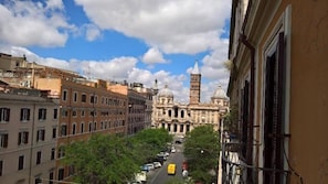 A street view with a historic building featuring a clock tower and domes, surrounded by multi-story buildings with balconies. - D'este (Rome)