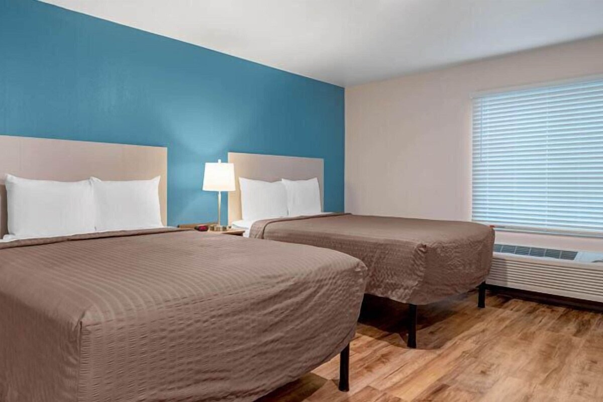 Una habitación de hotel con dos camas, una mesita de noche con lámpara, una ventana con persianas y un equipo de aire acondicionado.