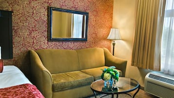 A hotel room with a sofa, a glass coffee table with a vase of flowers, a bed with a red blanket, a wall-mounted mirror, a lamp, and an air conditioning unit.