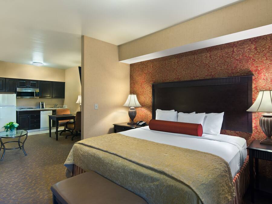 A hotel room with a bed, a desk, a chair, a lamp, a nightstand, and a kitchenette.