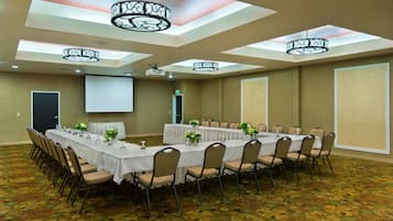 Un salón de conferencias con una larga mesa, sillas, una pantalla de proyector y luces en el techo.
