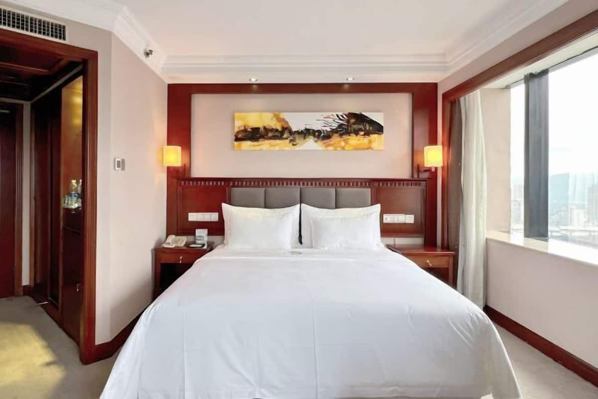 A hotel room with a large bed, a headboard with a cityscape painting, two bedside lamps, and a view of the city through a large window.