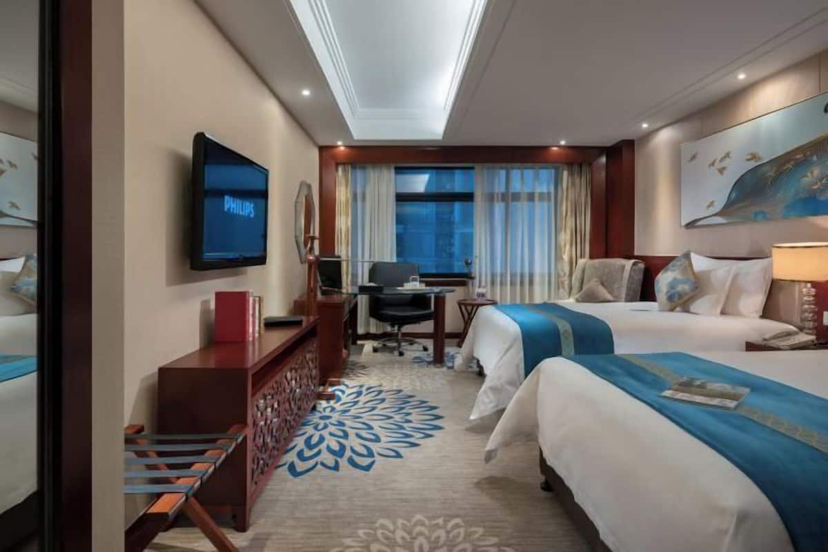 A hotel room with two beds, a desk, a chair, a television, and a large window with curtains.