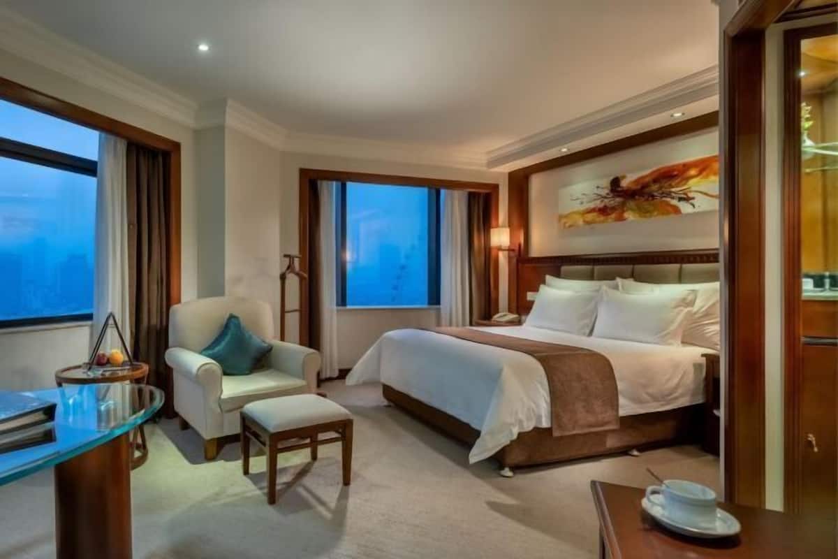 A hotel room with a bed, a desk, a chair, a window, and a painting on the wall.