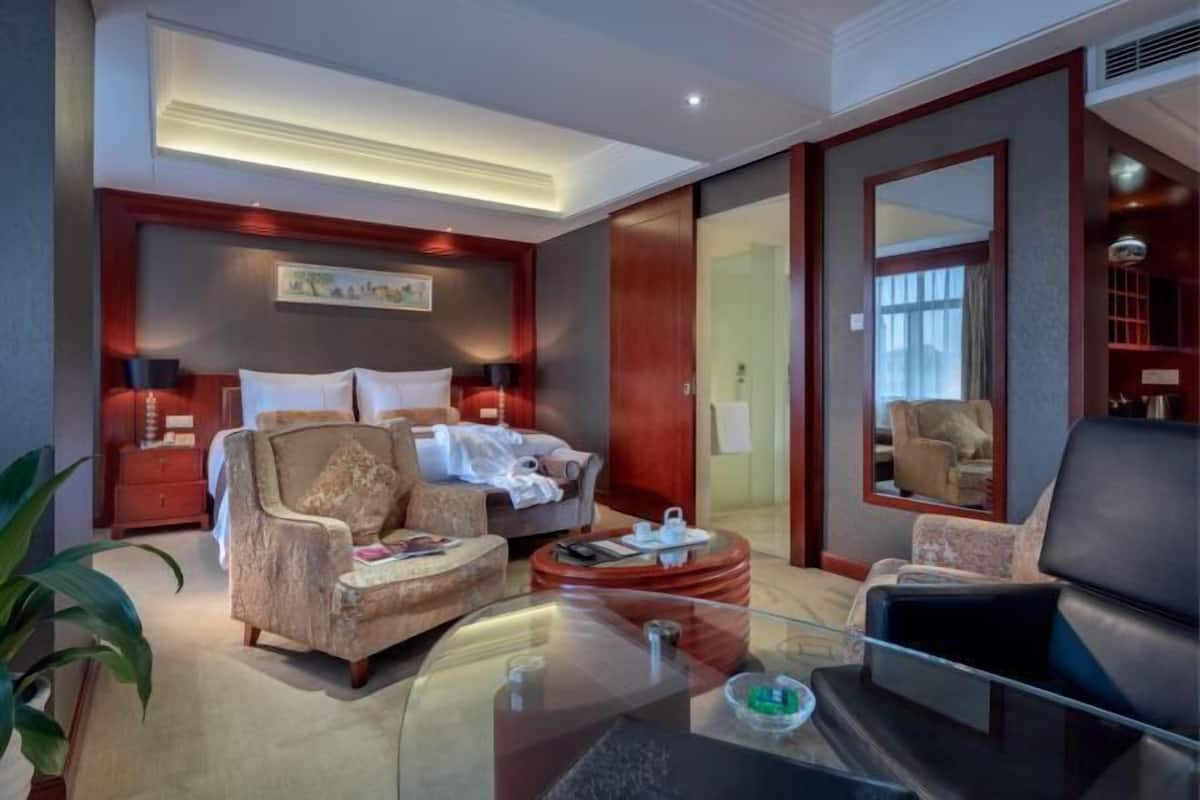 A hotel room with a bed, two armchairs, a glass coffee table, a side table, a mirror, and a large window.