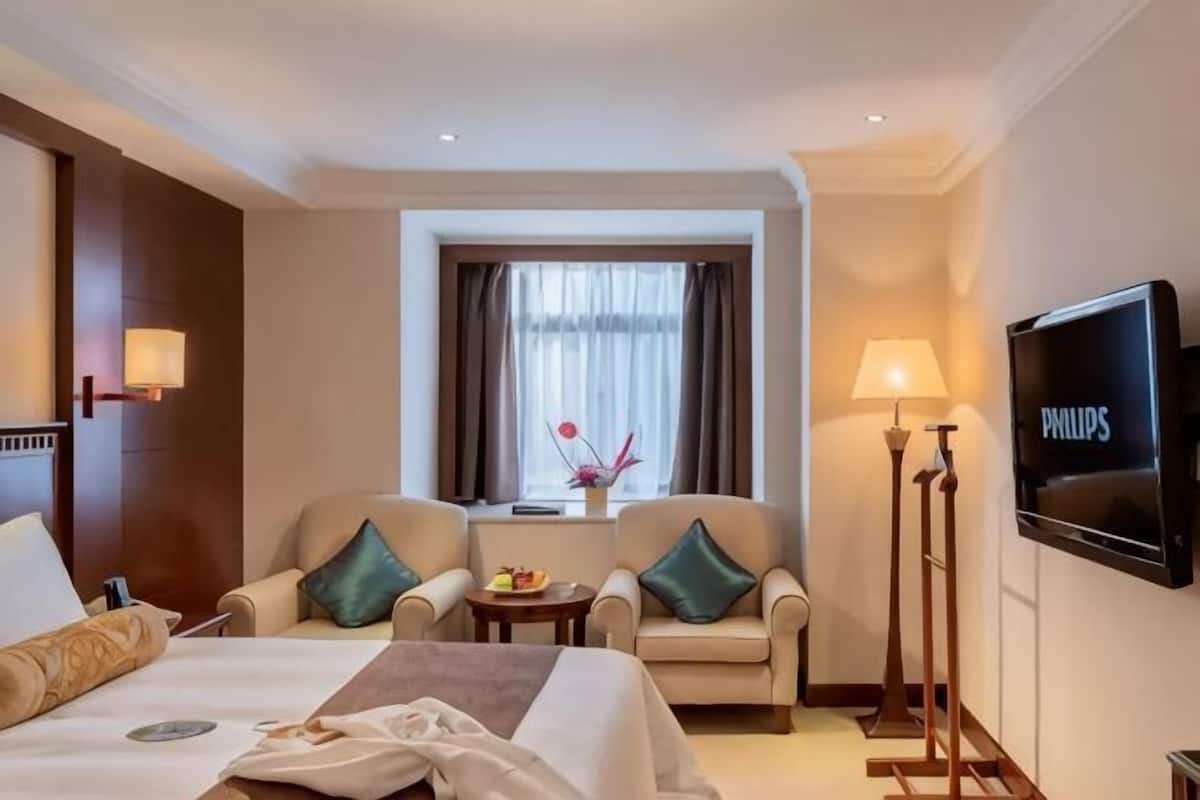 A hotel room with a bed, two armchairs, a small table with a fruit bowl, a window with curtains, a wall-mounted TV, and a lamp.