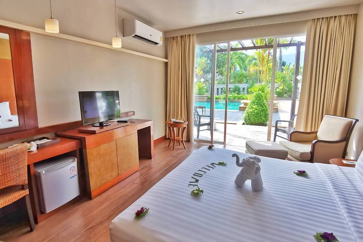 A hotel room with a bed, a desk with a television, a small refrigerator, and a view of a pool and greenery outside.