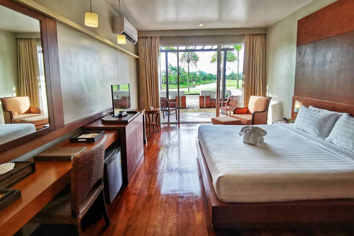 A hotel room with a large bed, a desk with a computer, a chair, and a view of a garden through large windows.
