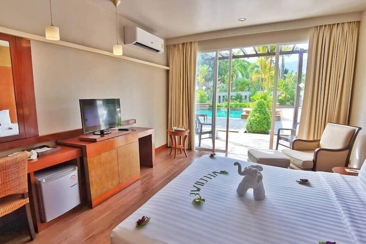 A hotel room with a bed, a desk with a TV, a small refrigerator, and a view of a pool and greenery.