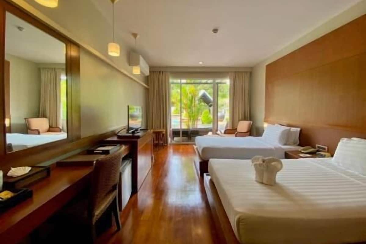 A hotel room with a large bed, a desk, a chair, a mirror, and a view of greenery outside.