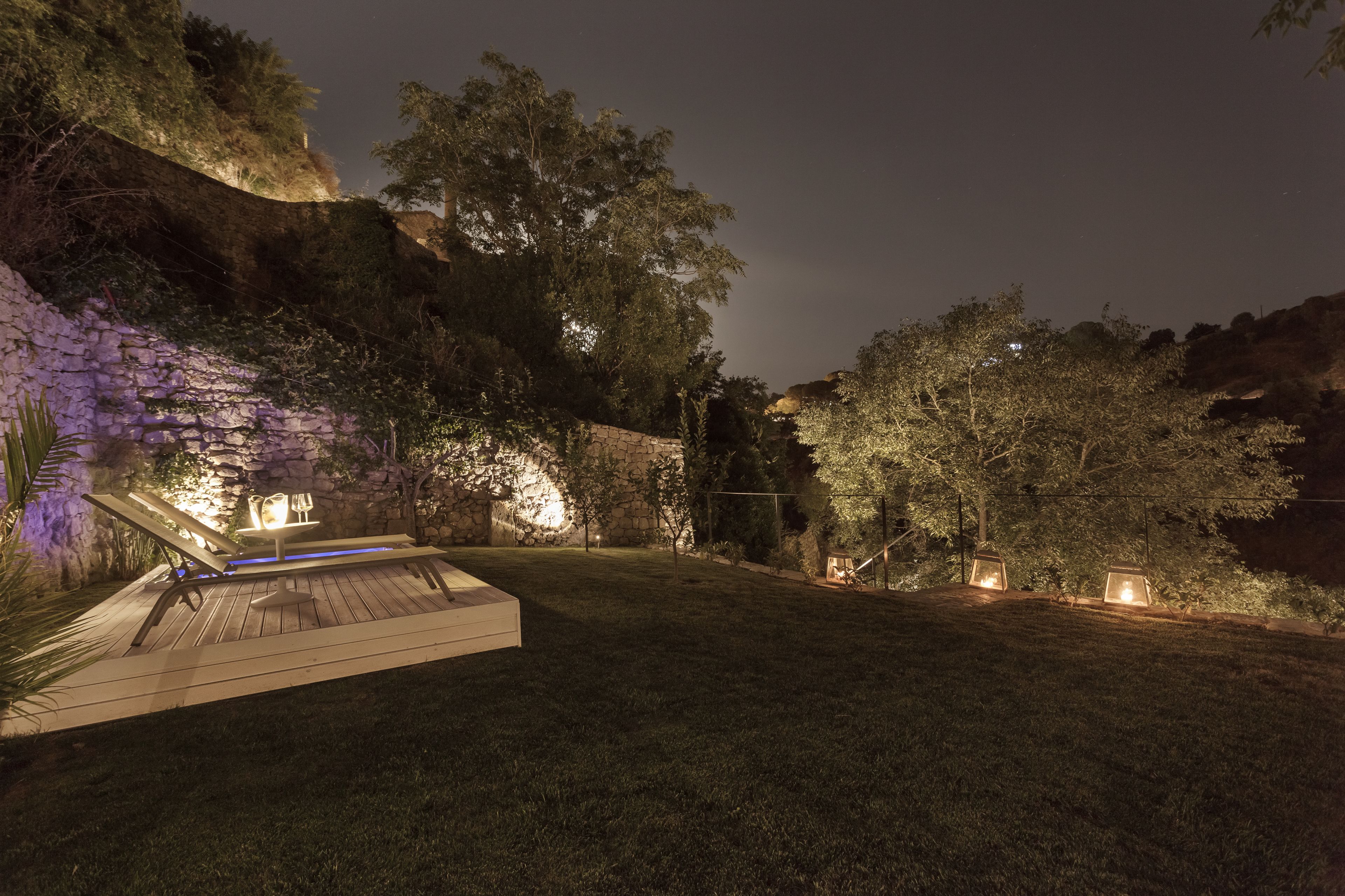 Un espace extérieur aménagé pour la nuit, avec une terrasse en bois, un mur en pierre et des arbres.