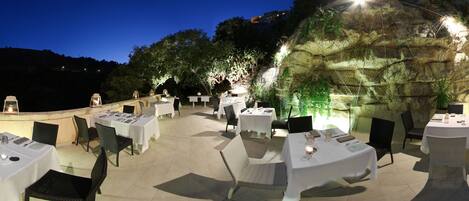 Un área de comedor al aire libre con mesas preparadas para dos, rodeada de sillas e iluminada con luz ambiental.