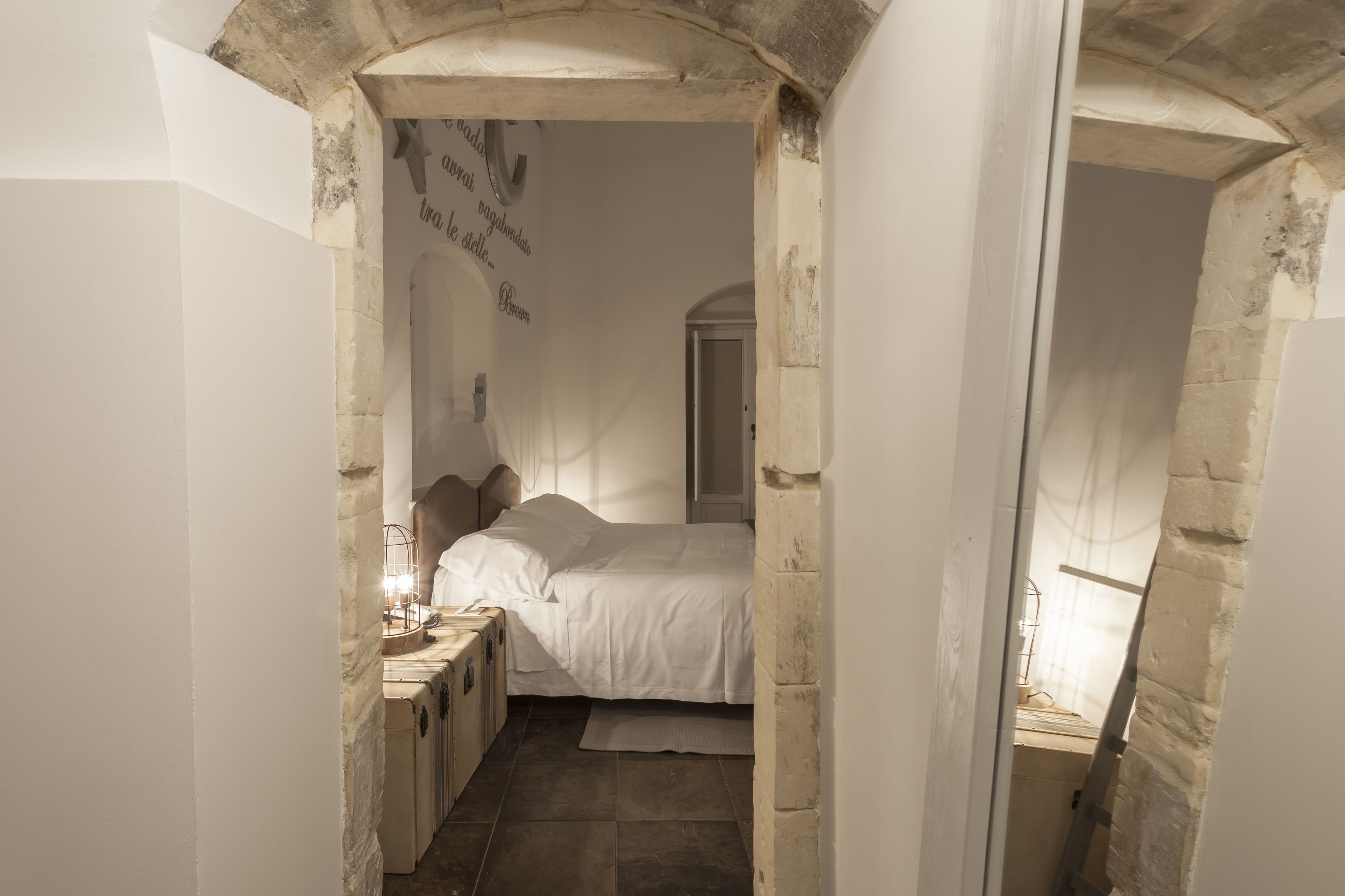 A narrow, stone-walled room with a bed, a nightstand, and a wall-mounted shelf.
