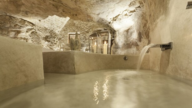Ein Thermalbad im Innenbereich mit Wasserspiel, Steinwänden und einer Kerzen-Nische.