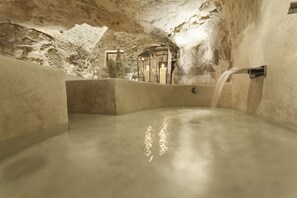 Une source thermale intérieure avec une fontaine à eau en mouvement, des murs en pierre et une alcôve éclairée aux chandelles.