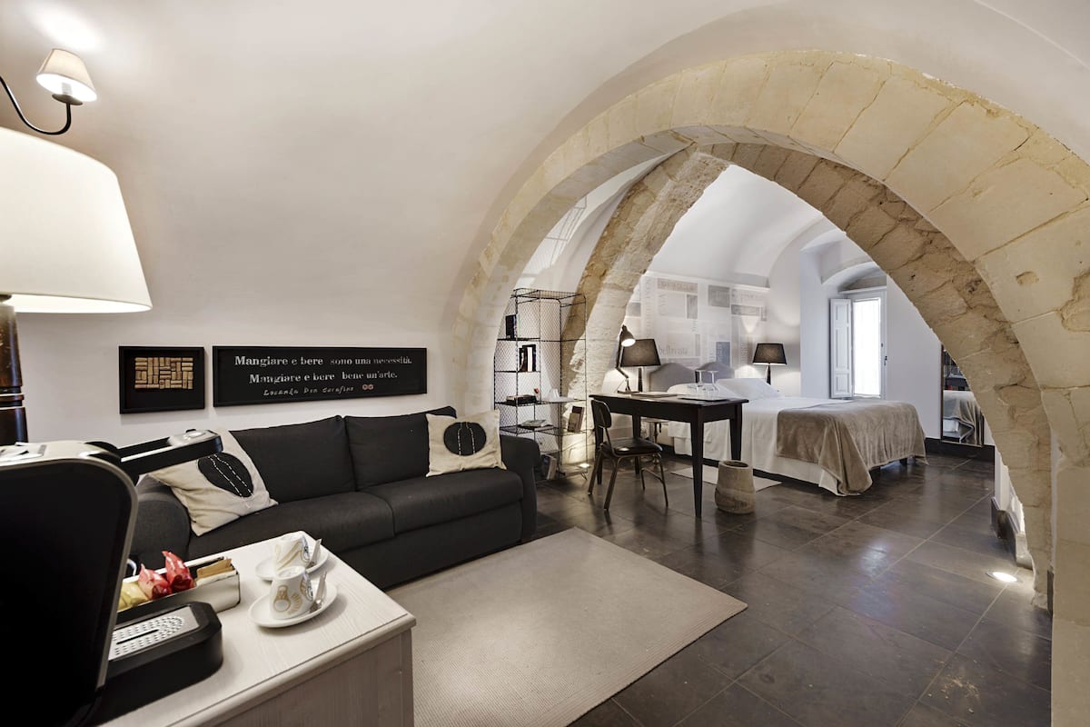 Ein modernes Hotelzimmer mit einer schwarzen Couch, einem Esstisch und einem Bett. Eine gebogene Steinarchitektur verbindet die beiden Räume.