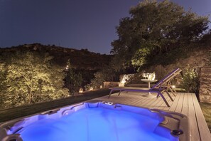 Un jacuzzi en una terraza de madera con una mesa y sillas, rodeado de árboles y un muro de piedra.