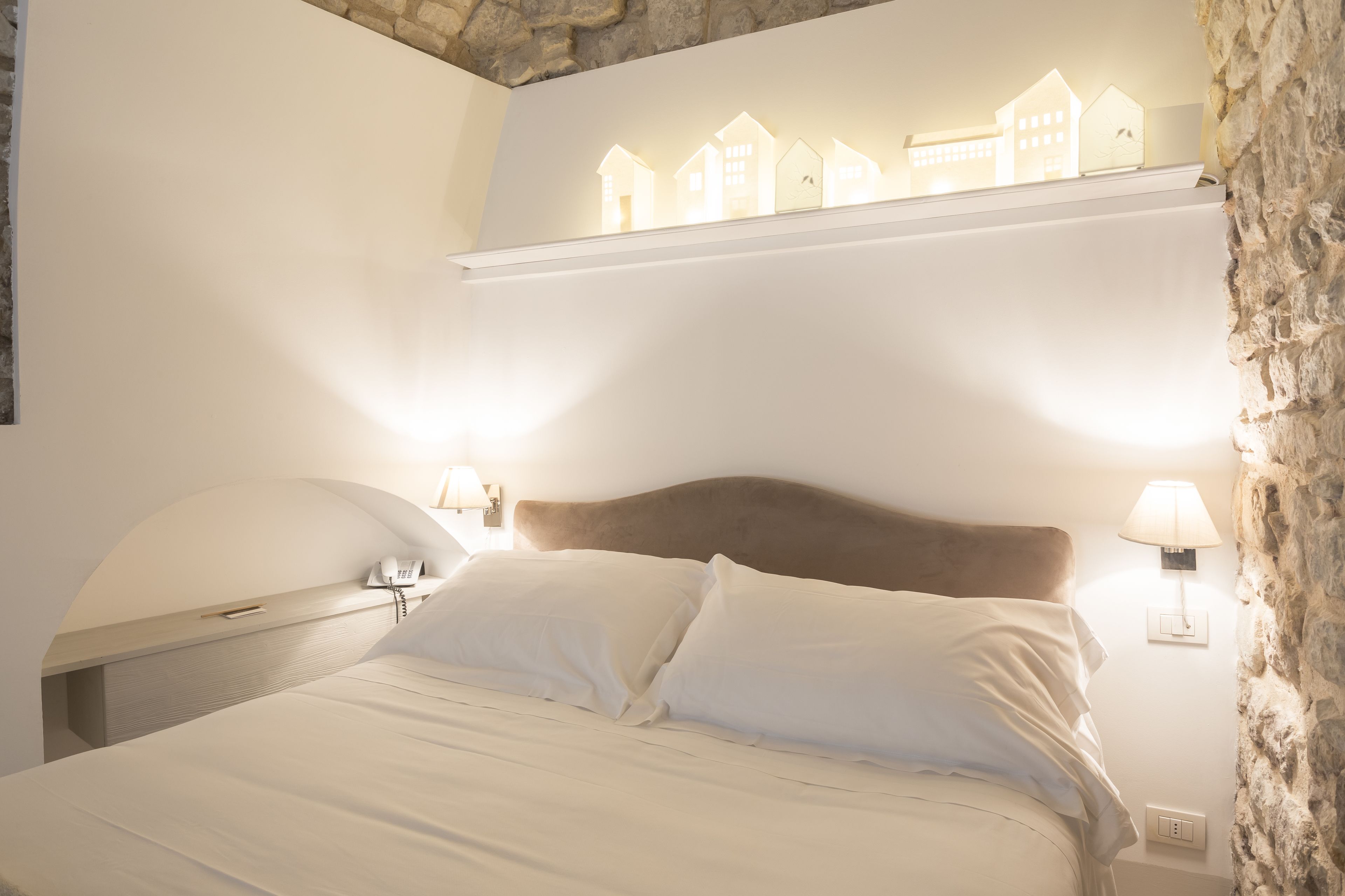 A bedroom with a bed, two bedside lamps, a nightstand, a wall-mounted lamp, and a shelf with house-shaped lights.