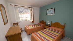 Une chambre avec deux lits simples, un bureau en bois, une chaise, une table de chevet, une lampe et une fenêtre avec des rideaux.