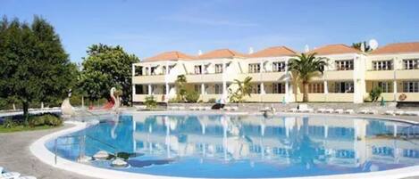 A swimming pool with lounge chairs, a building with a red roof, and greenery in the background.