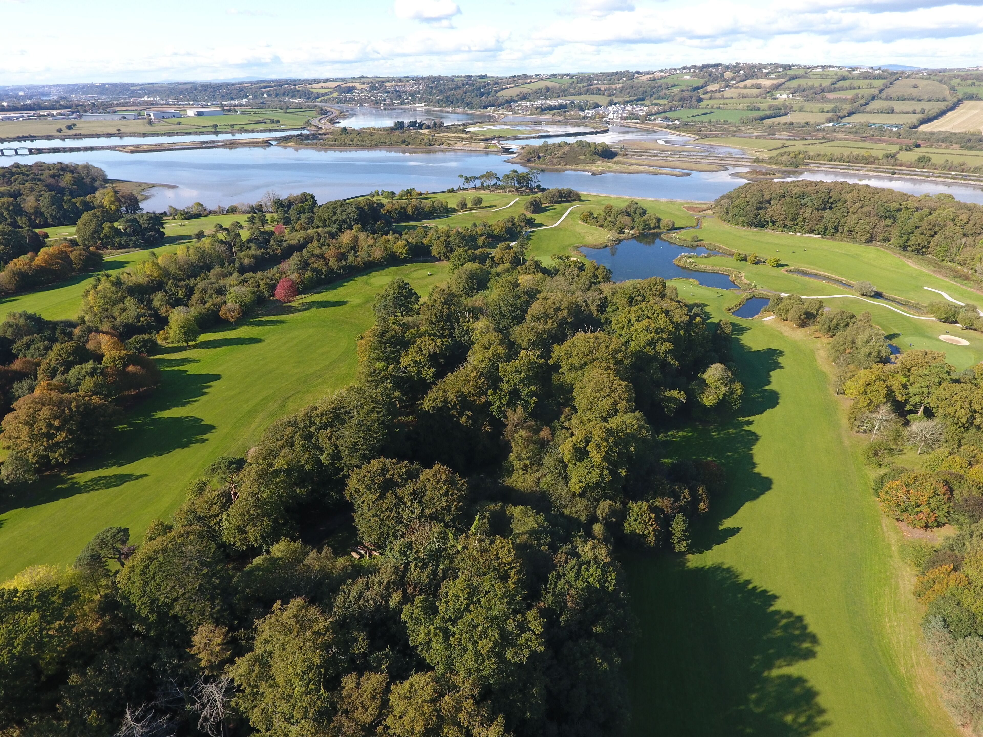 An aerial view of a golf course with multiple water bodies, surrounded by dense forests and residential areas in the background.