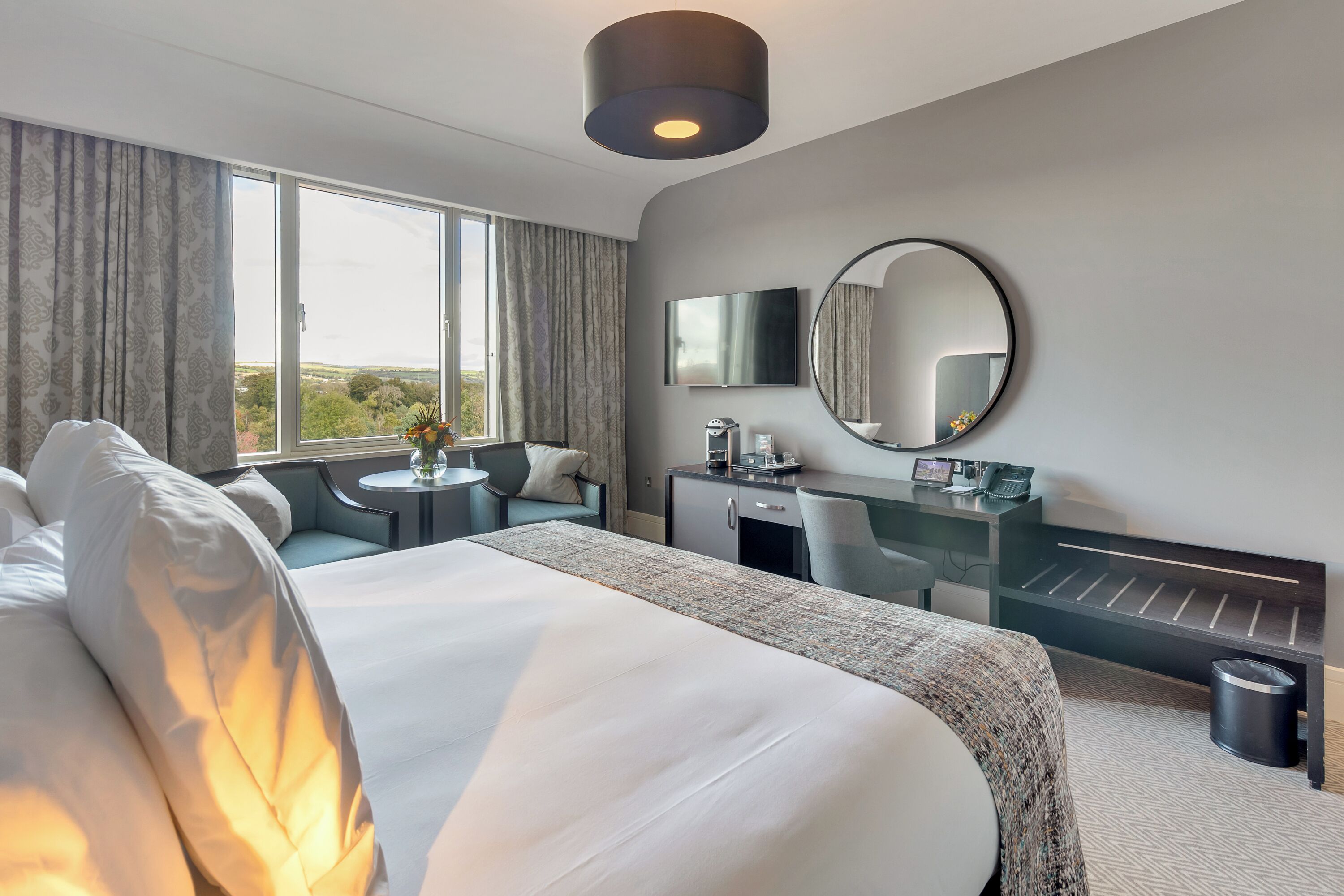 Una habitación de hotel moderna con una cama grande, un escritorio, un espejo redondo y vistas al exterior.