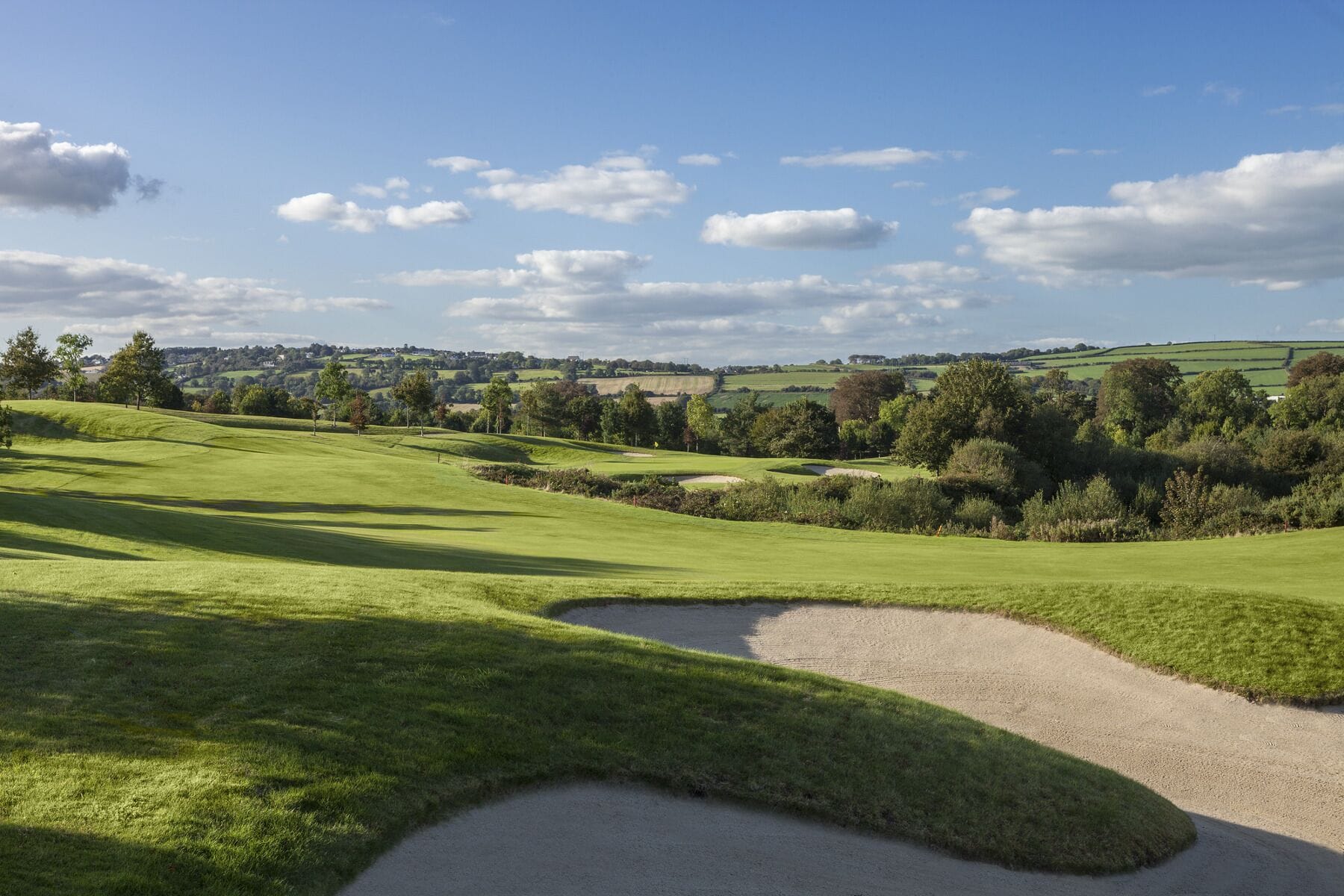 A golf course with rolling green hills, sand bunkers, and a clear sky.