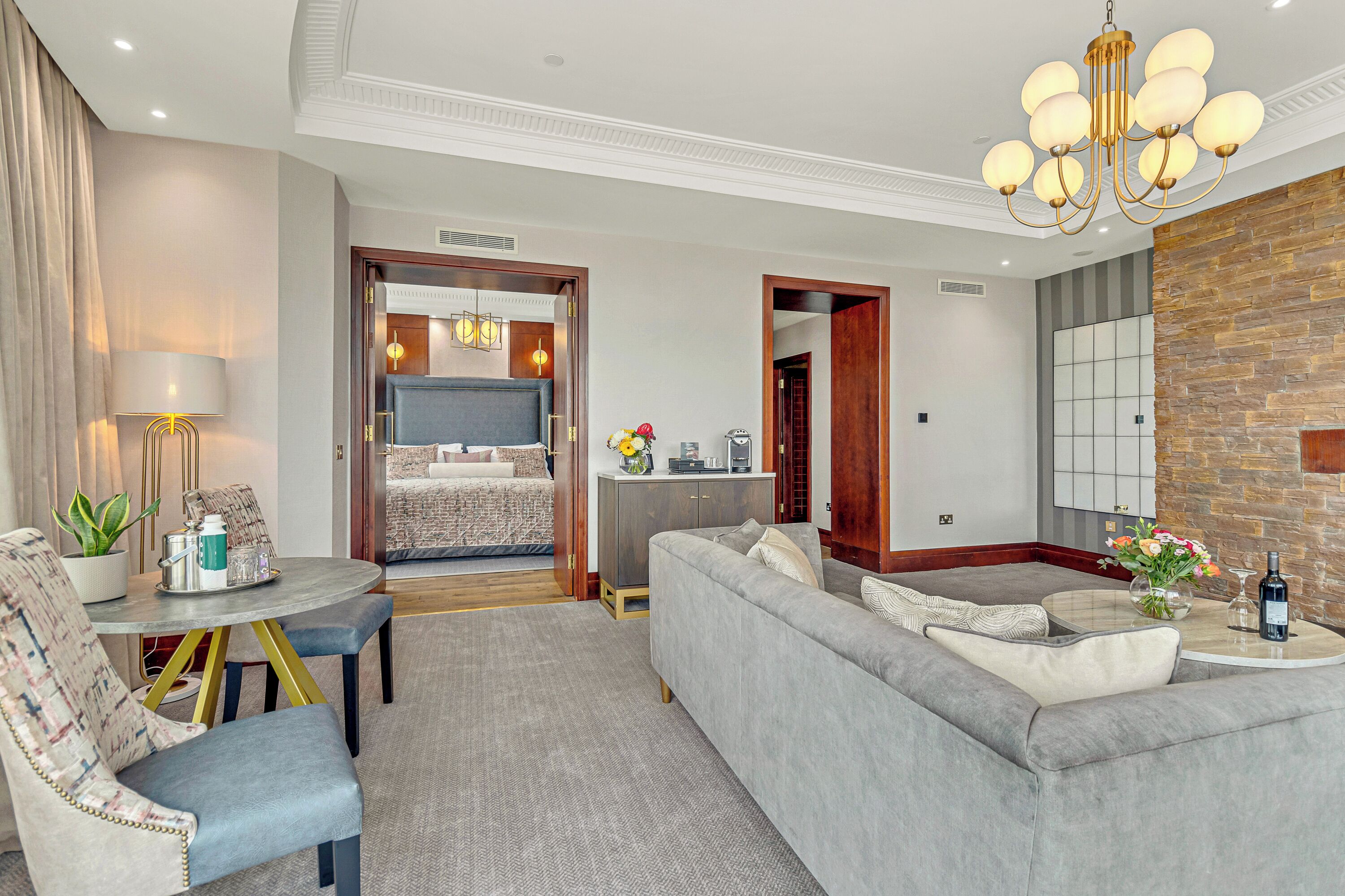 Un dormitorio de hotel moderno con un sofá gris, una mesa de comedor con sillas, una araña y vistas al resto del dormitorio a través de una puerta abierta.