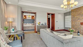 Un dormitorio de hotel moderno con un sofá gris, una mesa de comedor con sillas, una araña y vistas al resto del dormitorio a través de una puerta abierta.