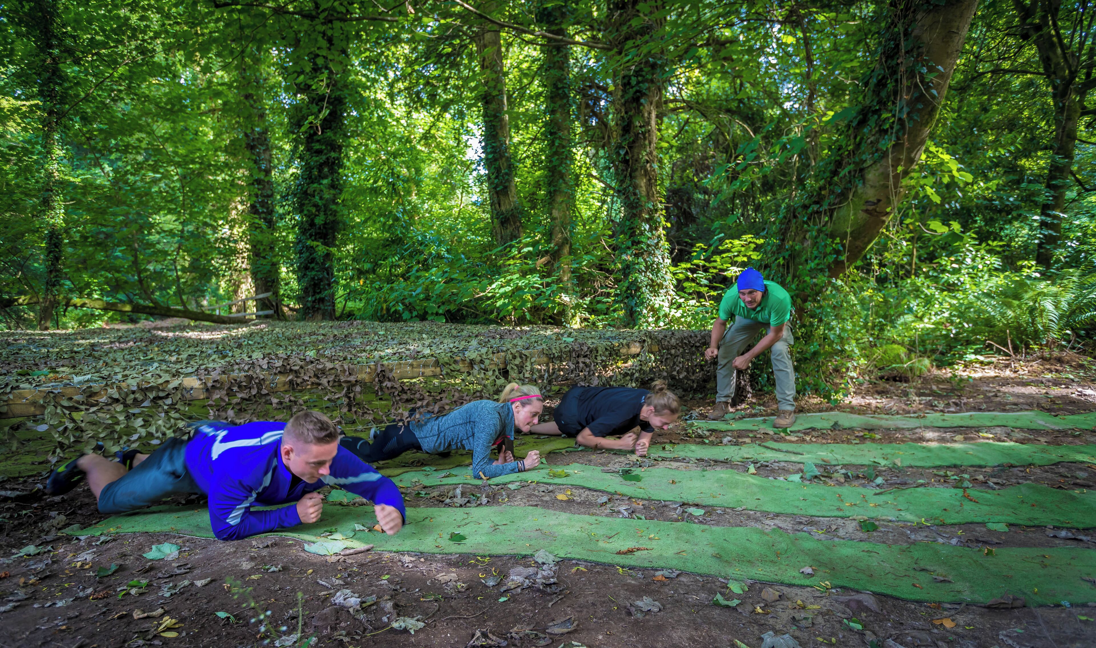 A group of people doing push-ups on green mats in a forested area with a stone wall in the background.