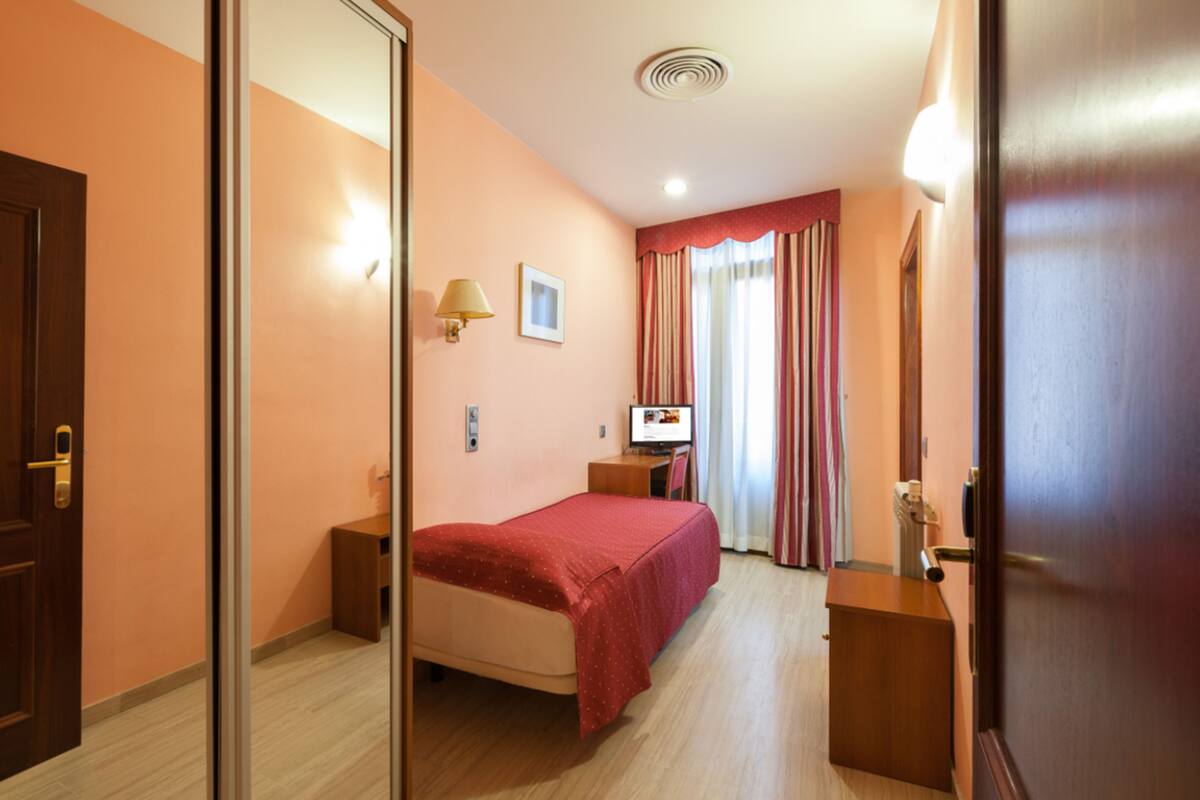A hotel room with a bed, a desk, a chair, a lamp, a TV, and a wardrobe.