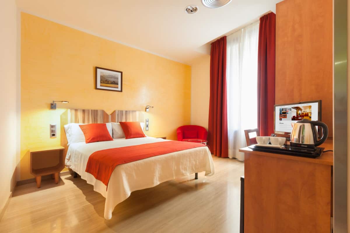A hotel room with a bed, bedside table, lamp, red chair, and a desk with a coffee maker.