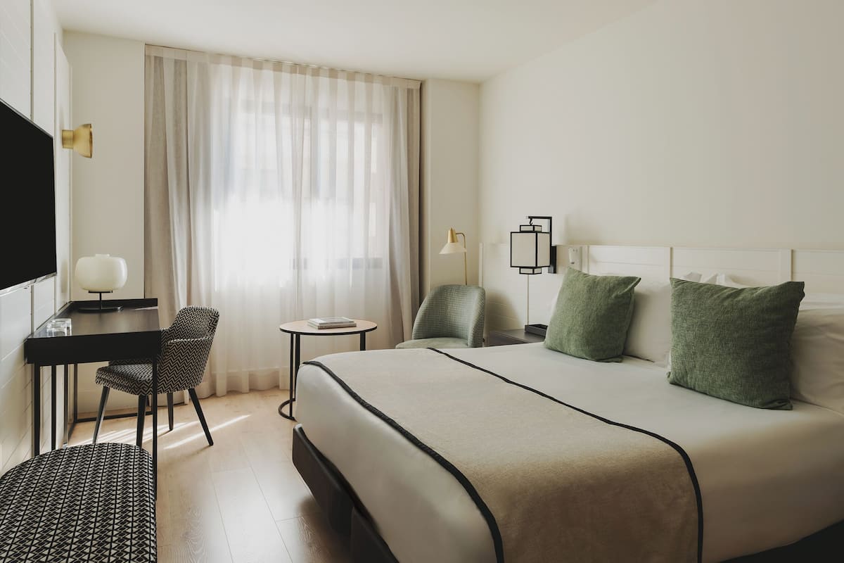 Habitación de hotel con cama, escritorio, silla, una mesita y ventana con cortinas.