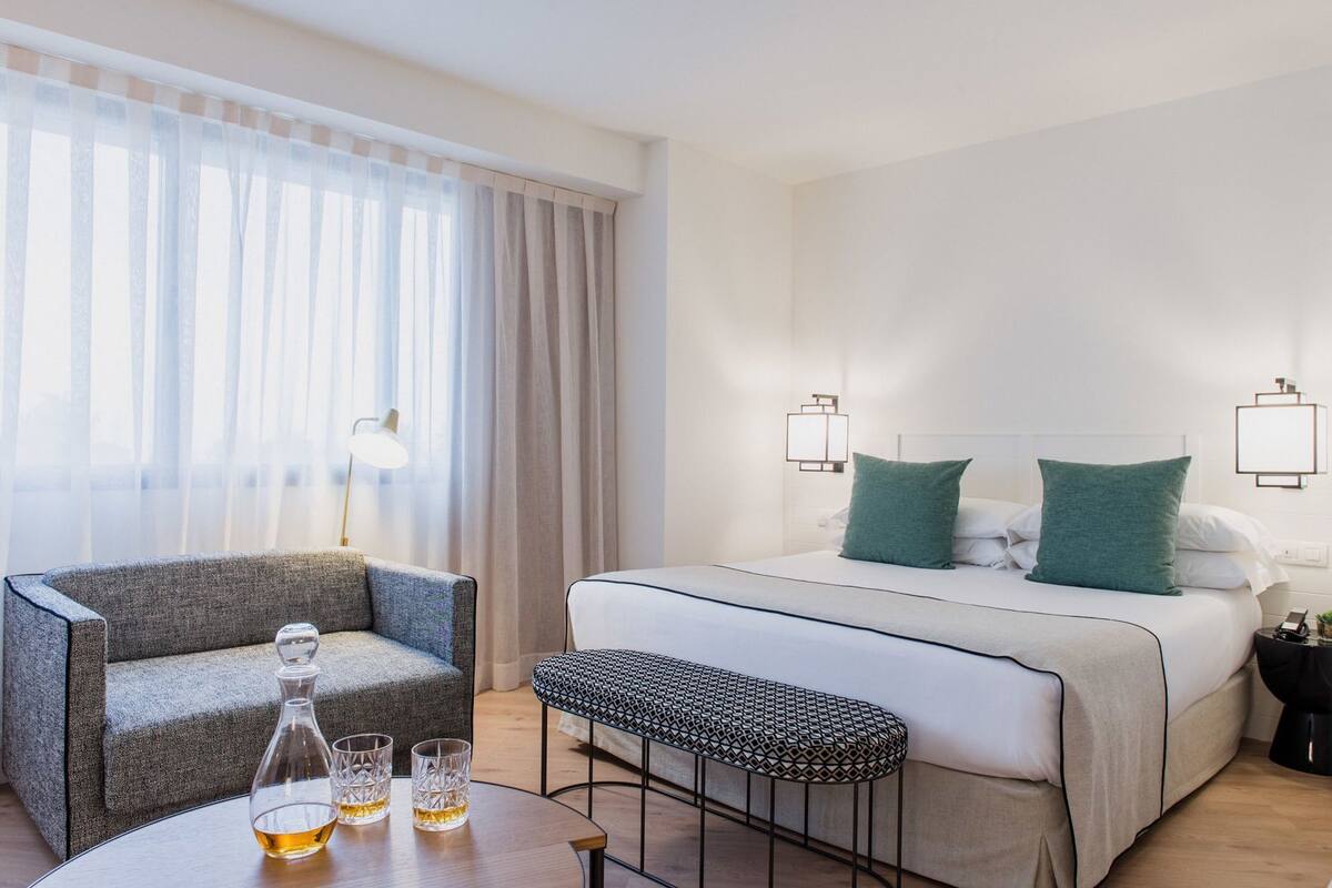 Una habitación de hotel con una cama, una silla, una mesita con vasos y una lámpara.