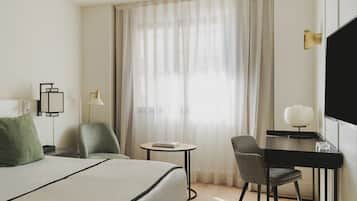 Et hotelværelse med seng, skrivebord, stol, fjernsyn og et vindue med gardiner.