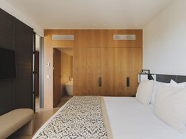 Un dormitorio moderno con una cama grande, un banco, armario de madera y un baño visible a través de una puerta abierta.