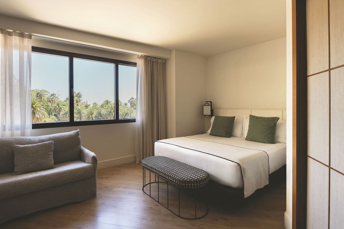 Una habitación de hotel con cama, sofá, banco y ventana con cortinas.