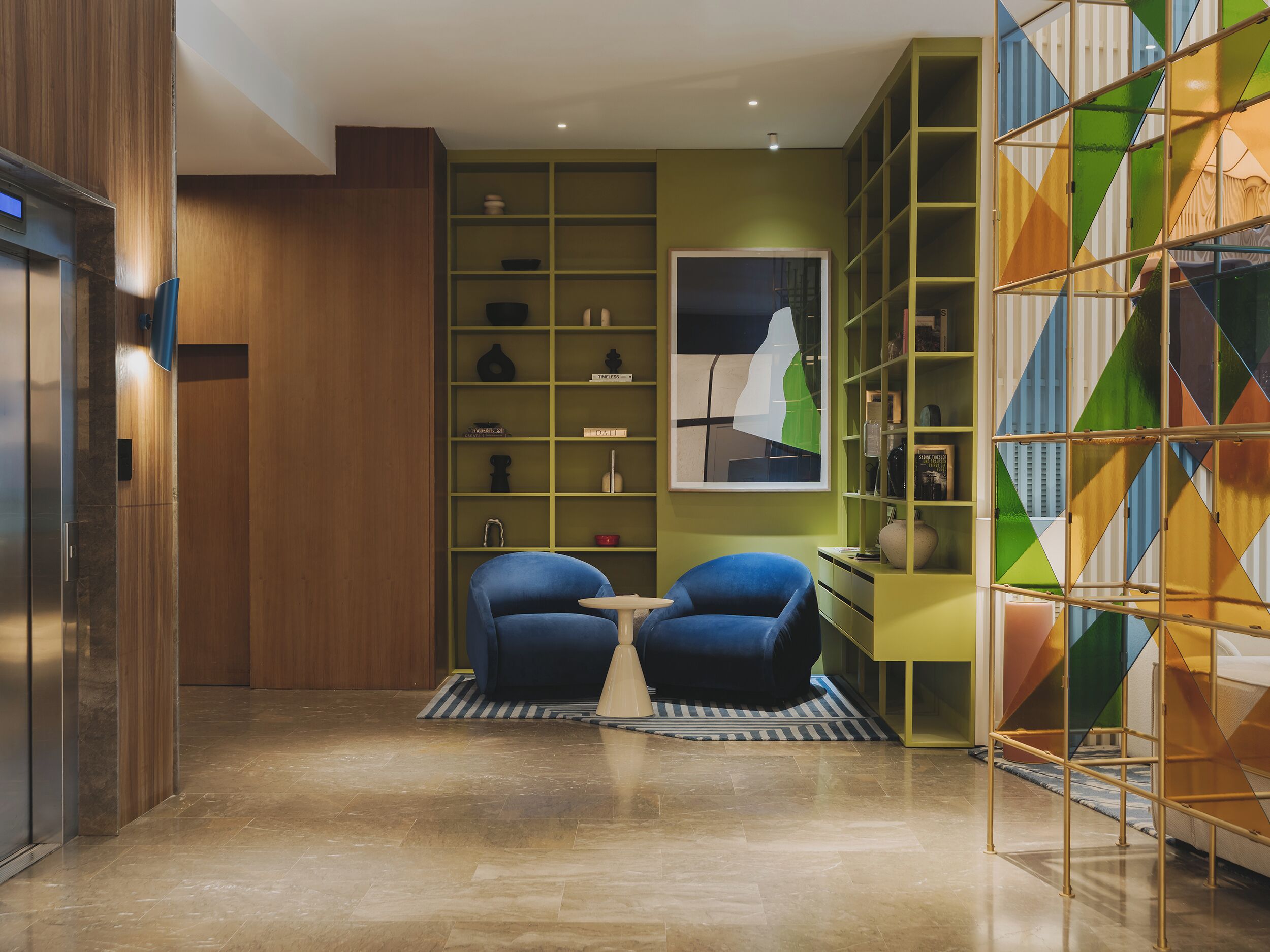 A modern interior with a wooden panel wall, a green shelving unit with various items, two blue armchairs, and a geometric-patterned glass partition.