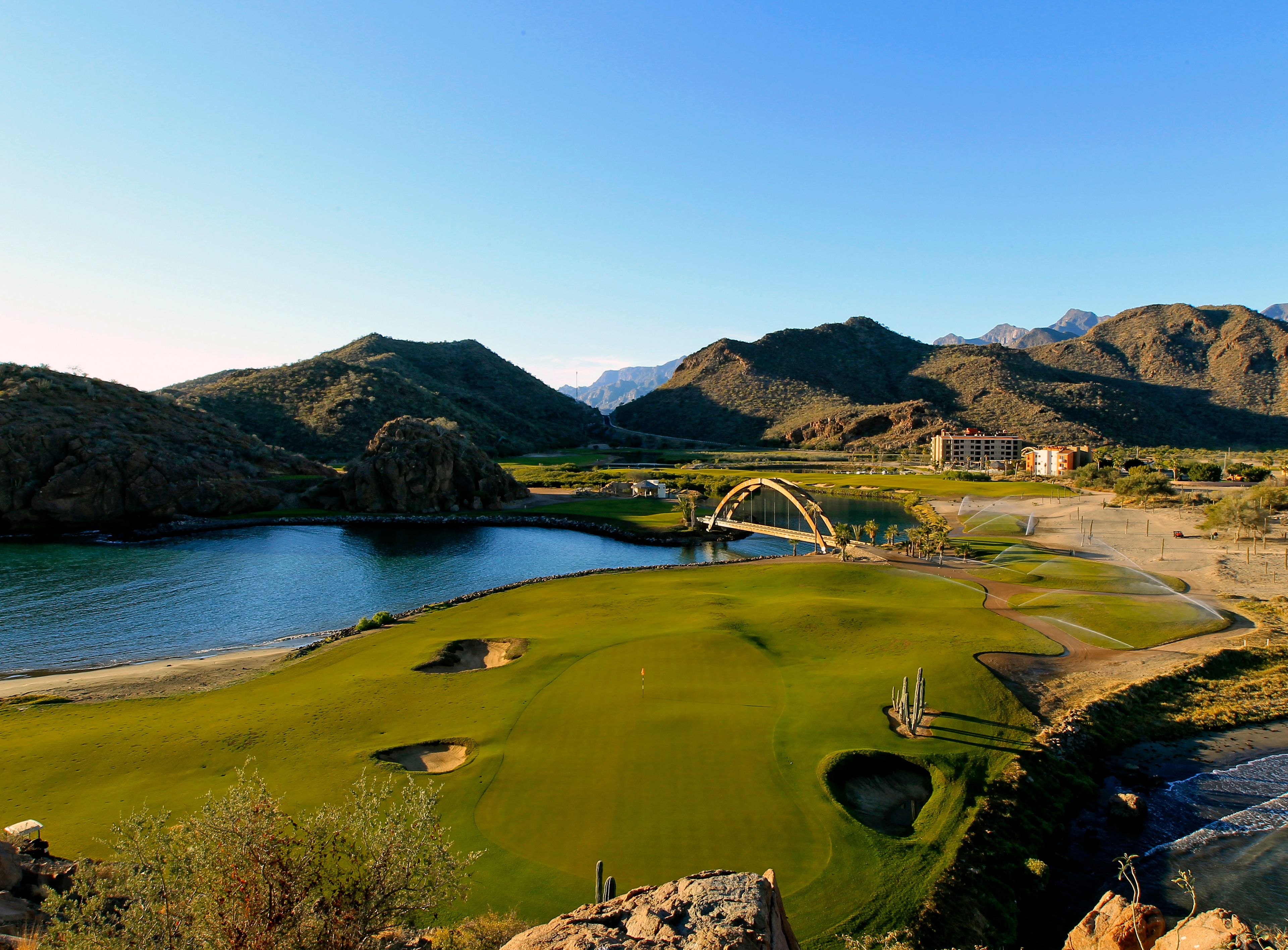 Un campo de golf con un puente, montañas y un cuerpo de agua.