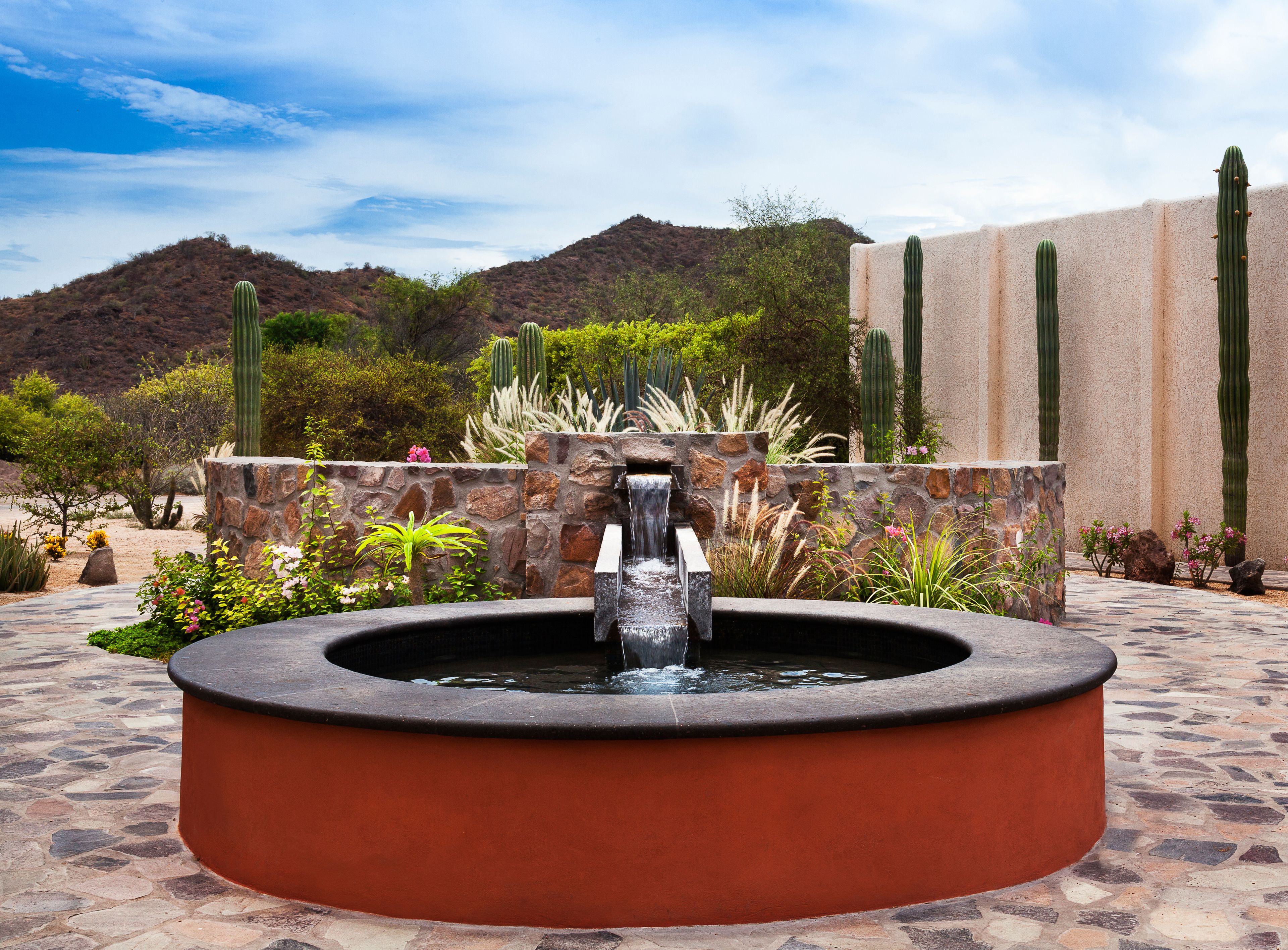 Una fuente de piedra circular con agua brotando del centro, rodeada de diversas plantas desérticas y cactus.