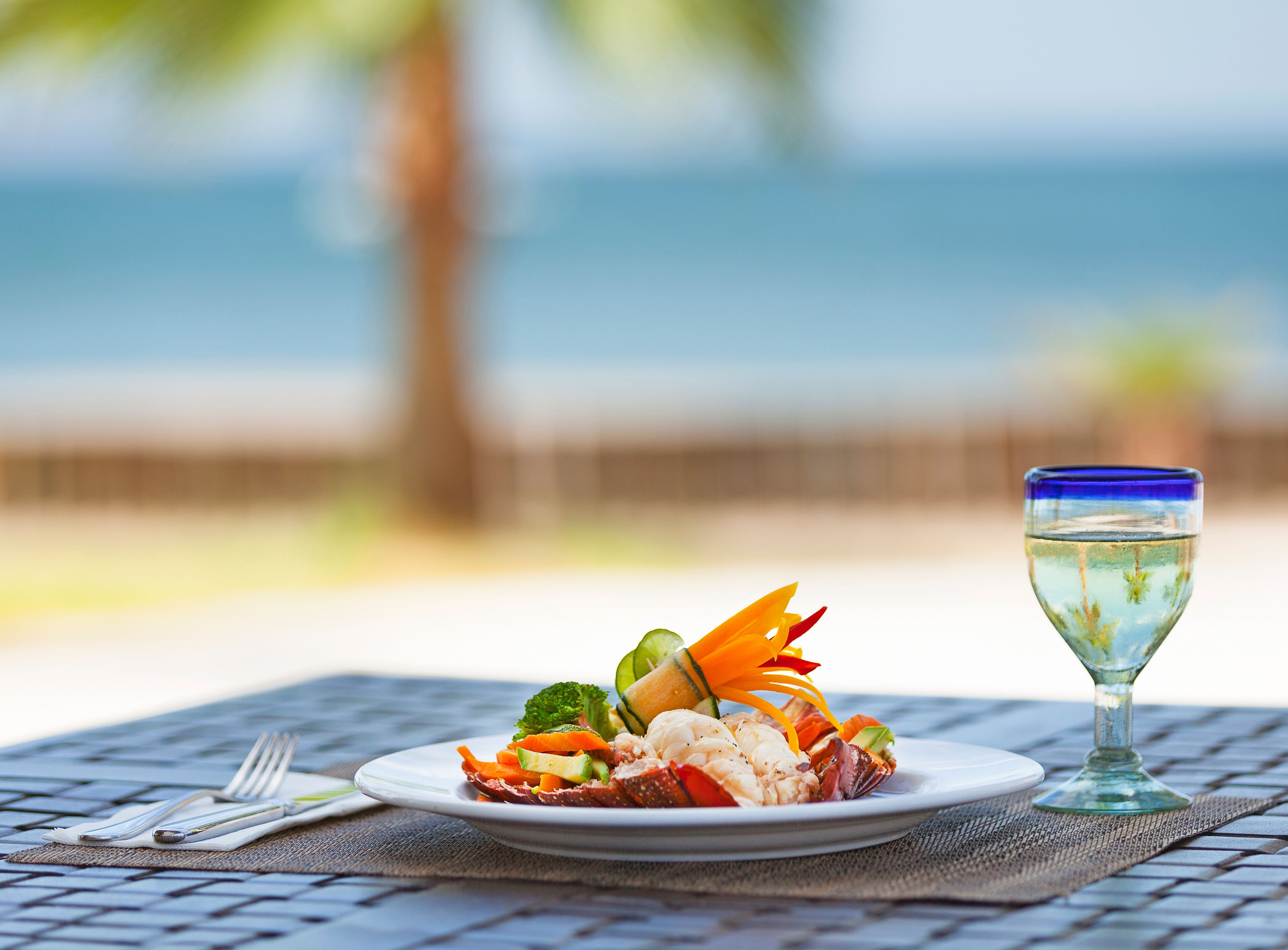 A plate of seafood and vegetables on a table with a glass of water, set against a blurred beach and ocean background.