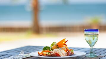 A plate of seafood and vegetables on a table with a glass of water, set against a blurred beach and ocean background.