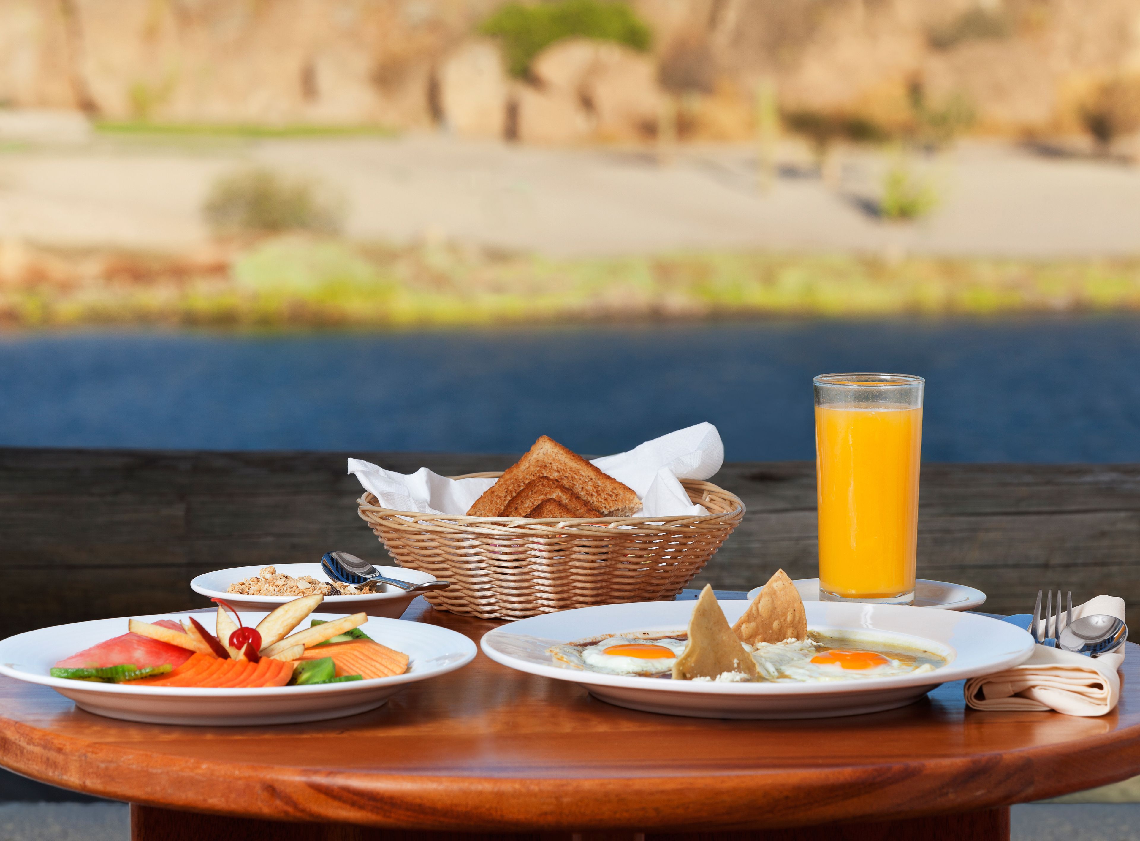 Un desayuno con platos de comida, una cesta de pan y un vaso de jugo de naranja sobre una mesa de madera, con vistas a una masa de agua al fondo.