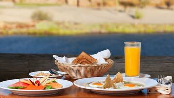 Disposição de café da manhã com pratos de comida, cesta de pão e copo de suco de laranja sobre uma mesa de madeira, com vista para um corpo d'água ao fundo.