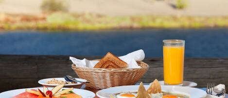 Un desayuno con platos de comida, una cesta de pan y un vaso de jugo de naranja sobre una mesa de madera, con vistas a una masa de agua al fondo.