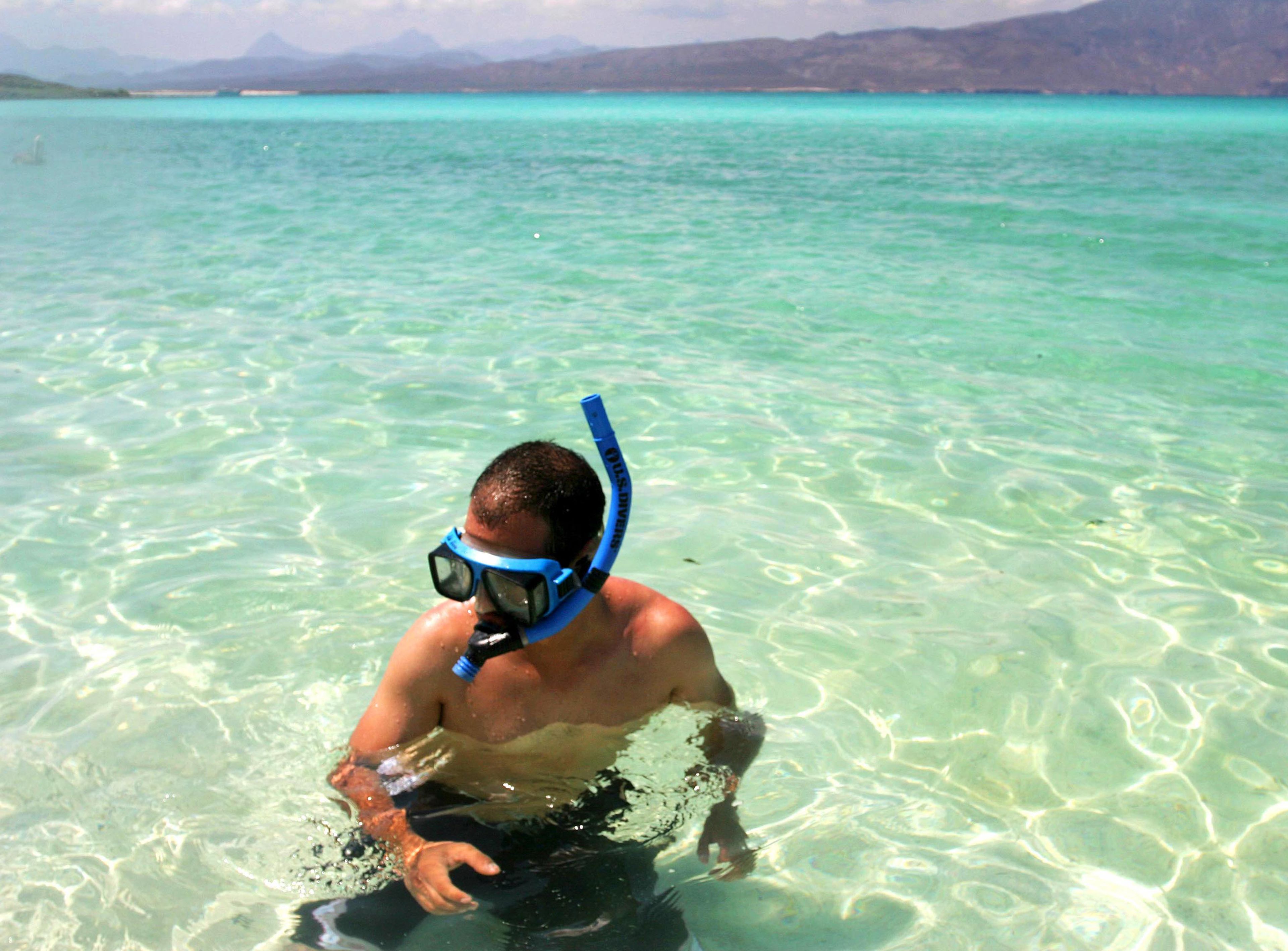 A person snorkeling in clear turquoise water with mountains in the background.