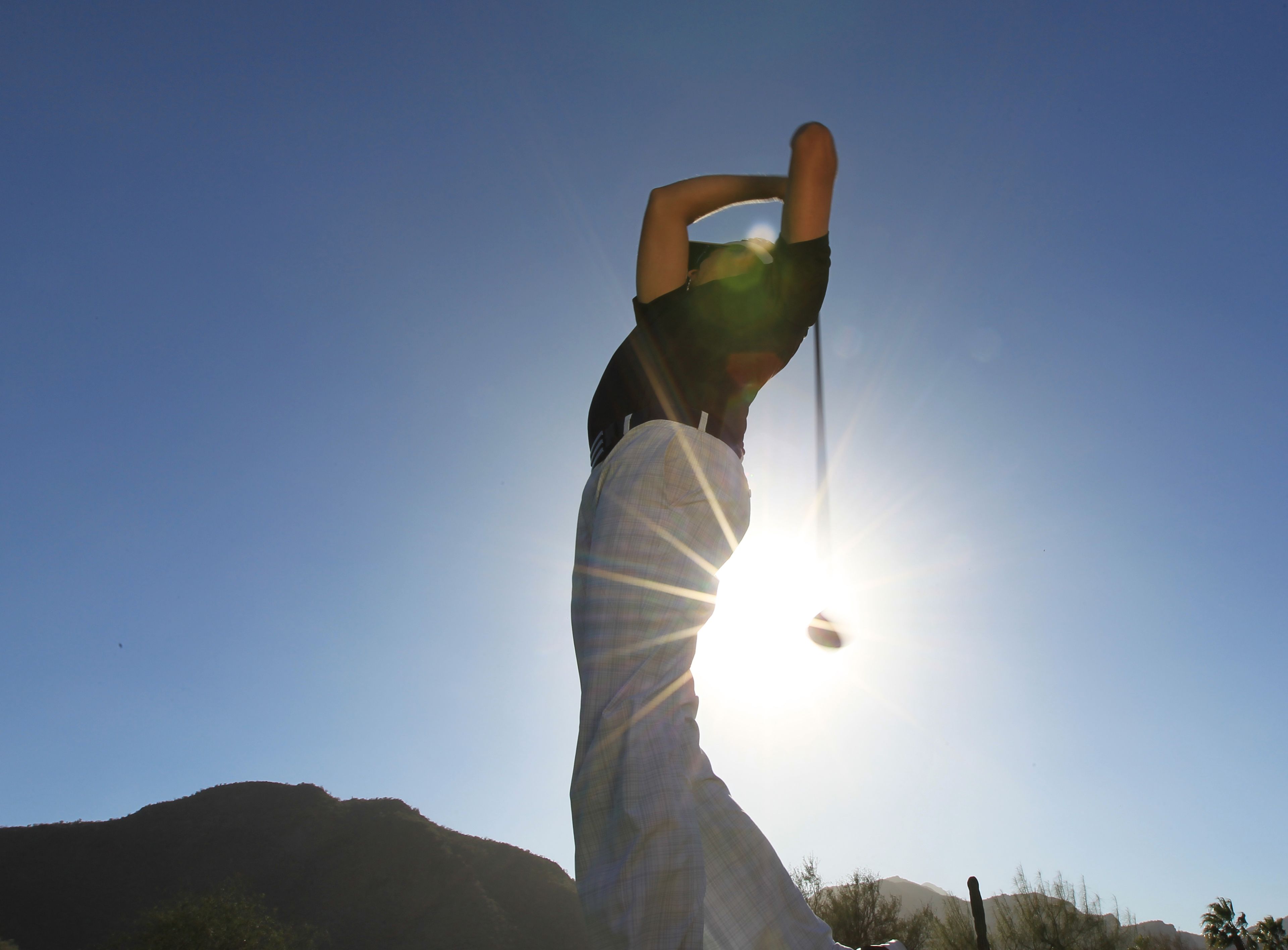 A person in a plaid shirt and shorts, holding a golf club, with the sun in the background.
