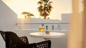 A round table with a glass of amber liquid and a bowl of nuts, a black chair, and a view of palm trees and buildings at sunset. - Coral California (Arona)