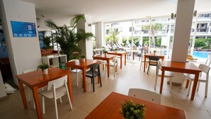A modern indoor dining area with wooden tables and chairs, potted plants, and a view of a swimming pool and buildings outside. - Coral California (Arona)